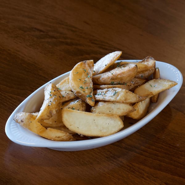 A white oval dish filled with golden-brown potato wedges, seasoned with herbs and salt, on a wooden table.