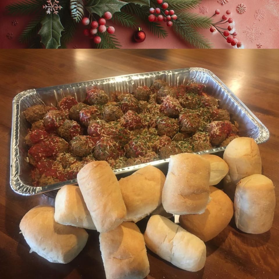 A catering tray of meatballs with sauce and cheese, surrounded by dinner rolls, on a wooden table.